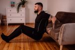 A bearded man in black fitness attire exercising at home, showcasing arm tattoos and a healthy lifestyle.