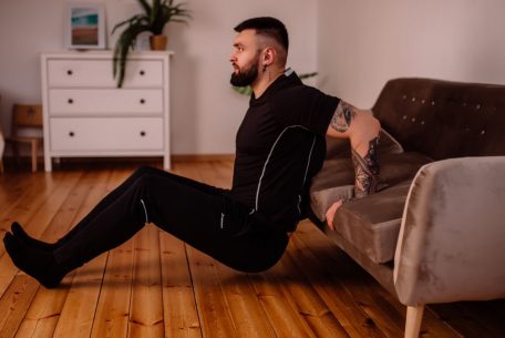 A bearded man in black fitness attire exercising at home, showcasing arm tattoos and a healthy lifestyle.